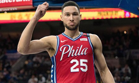 Ben Simmons in a red Philadelphia 76ers jersey holding his arm up celebrating after scoring, looking right into the camera.