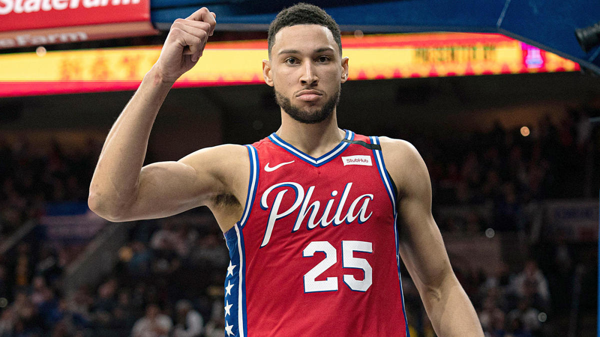 Ben Simmons in a red Philadelphia 76ers jersey holding his arm up celebrating after scoring, looking right into the camera.