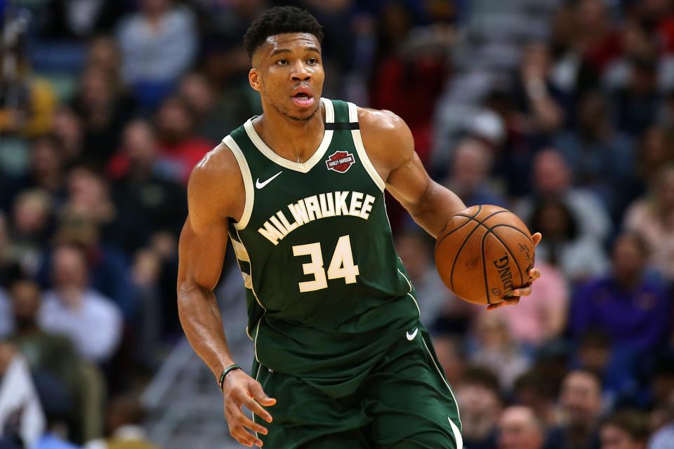 Giannis Antetokounmpo dribbling a basketball, wearing the green Milwaukee Bucks uniform in front of a crowd from 2019.